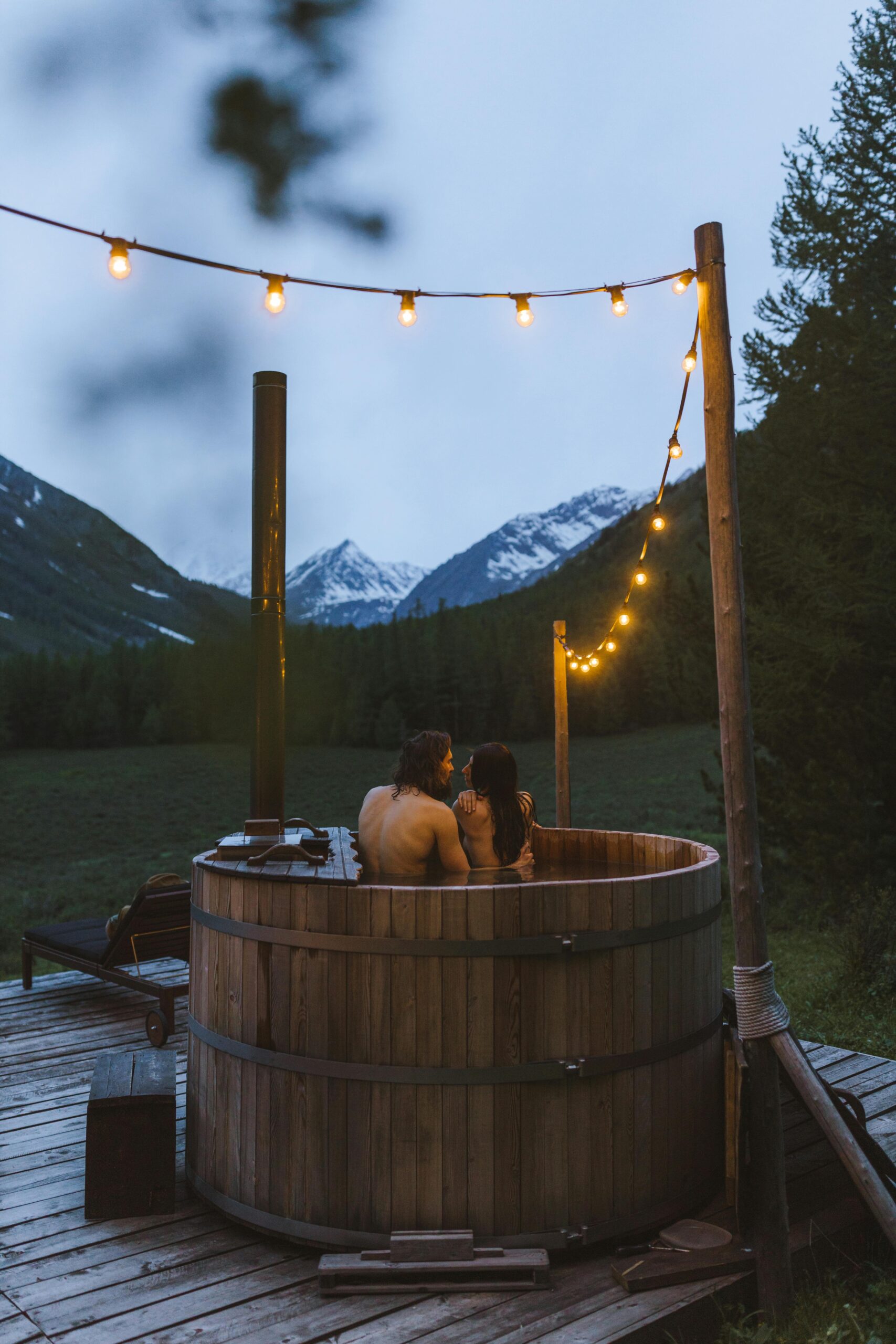 Couple relaxing in an outdoor wooden hot tub with mountain views and string lights at dusk.